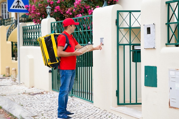 Vista lateral del mensajero caucásico que suena en el timbre. Cartero pensativo con uniforme rojo, llevando una bolsa térmica amarilla, sosteniendo el paquete y de pie al aire libre. Servicio de entrega y concepto de correo.