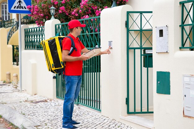 Vista lateral del mensajero caucásico que suena en el timbre. Cartero pensativo con uniforme rojo, llevando una bolsa térmica amarilla, sosteniendo el paquete y de pie al aire libre. Servicio de entrega y concepto de correo.