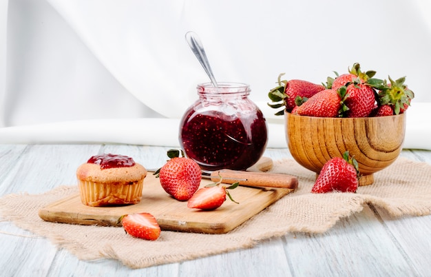 Vista lateral mermelada de fresa y cupcake con fresa fresca en un tazón de madera y un cuchillo sobre fondo blanco.