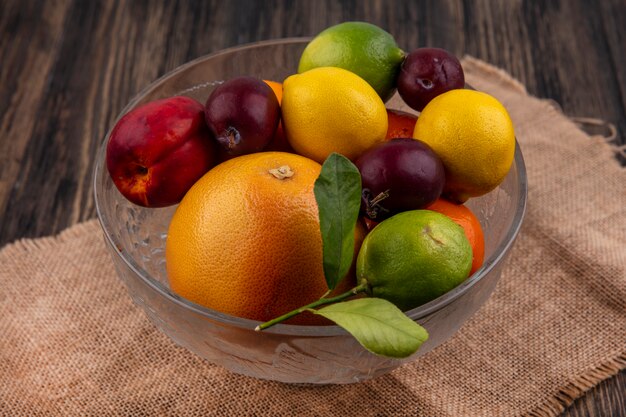 Vista lateral de la mezcla de frutas limones limas ciruelas melocotones y naranjas en un jarrón sobre una servilleta beige