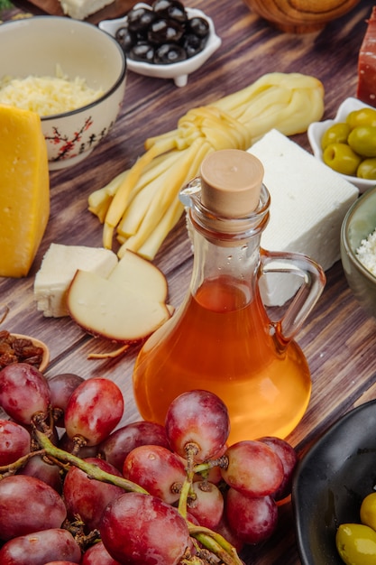 Vista lateral de la miel en una botella de vidrio con aceitunas en vinagre uvas frescas y varios tipos de queso en madera rústica