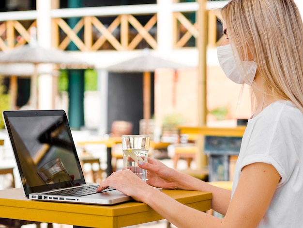 Vista lateral de la mujer al aire libre con mascarilla trabajando en la computadora portátil