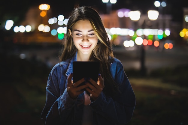 Vista lateral de la mujer atractiva con tableta digital en las calles de la ciudad por la noche