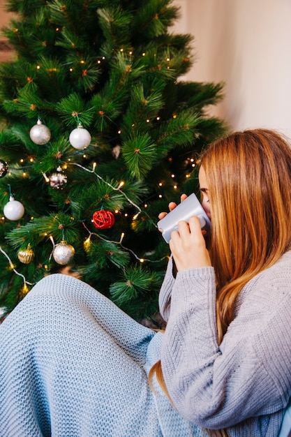 Vista lateral de mujer bebiendo té enfrente de árbol de navidad