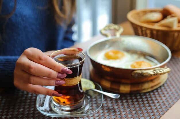 Vista lateral mujer bebiendo té con huevos fritos en una sartén con pan en una cesta