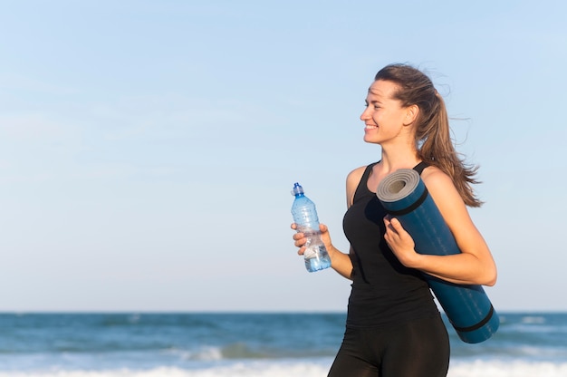 Foto gratuita vista lateral de la mujer con botella de agua y esterilla de yoga en la playa