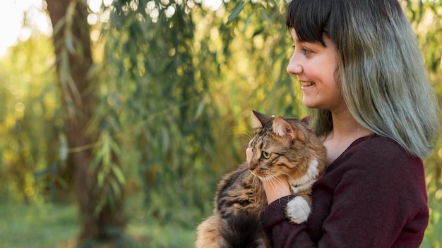 Vista lateral de una mujer de cabello teñido abrazando a su gato atigrado en el bosque