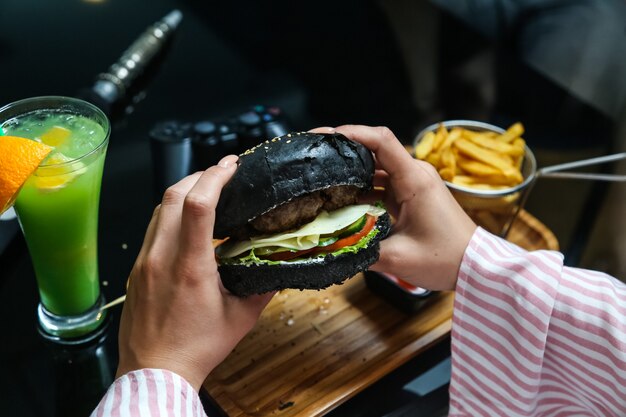 Vista lateral mujer come hamburguesa negra con papas fritas y salsa de tomate con mayonesa en un stand con un cóctel