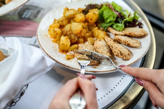 Vista lateral mujer come nuggets de pollo con papas y hojas de ensalada en un plato