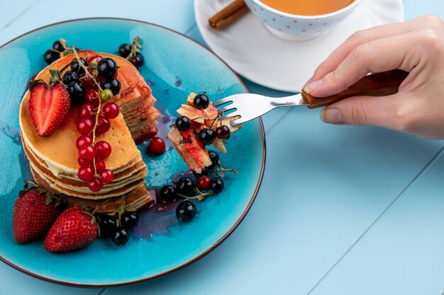 Vista lateral de una mujer come panqueques con fresas, grosellas rojas y negras y una taza de té sobre una superficie azul