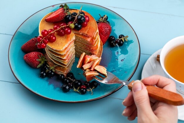 Vista lateral de una mujer come panqueques con fresas, grosellas rojas y negras y una taza de té sobre una superficie azul