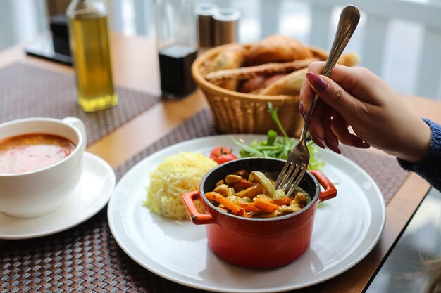 Vista lateral mujer comiendo pollo con verduras en una cacerola con arroz y hierbas con tomate en un plato