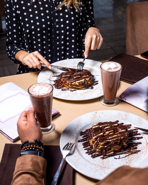 Vista lateral de una mujer comiendo postre con plátanos cubiertos de chocolate y servido con cacao con malvavisco en vidrio en la mesa