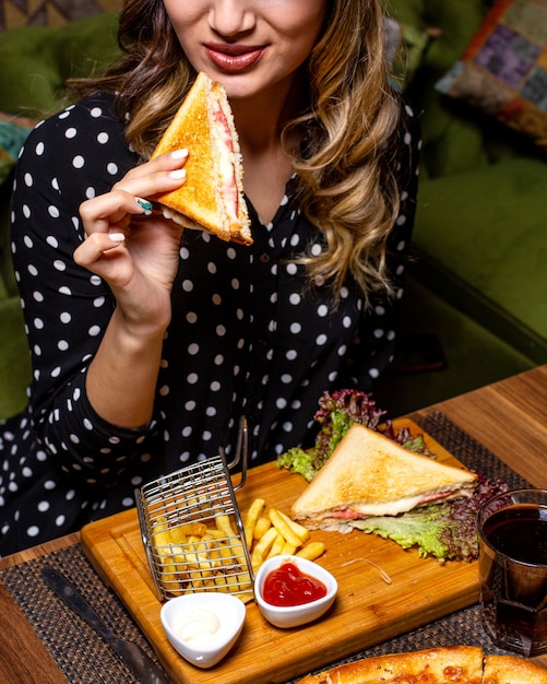 Vista lateral de una mujer comiendo sándwich club servido con papas fritas y salsa de tomate en la mesa