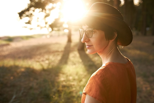Vista lateral de una mujer elegante y pensativa con el pelo corto que camina al aire libre, con gafas y sombrero, disfrutando de una agradable velada con rayos de luz solar a través de las hojas de los árboles.