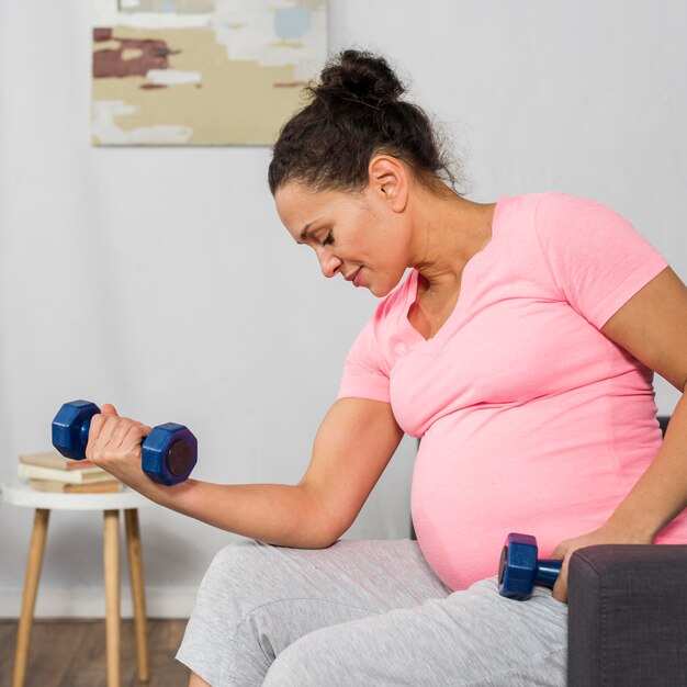 Vista lateral de la mujer embarazada en casa haciendo ejercicio con pesas