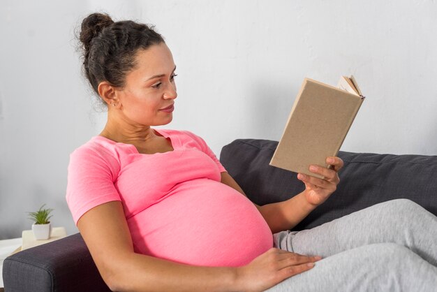 Vista lateral de la mujer embarazada en casa leyendo en el sofá