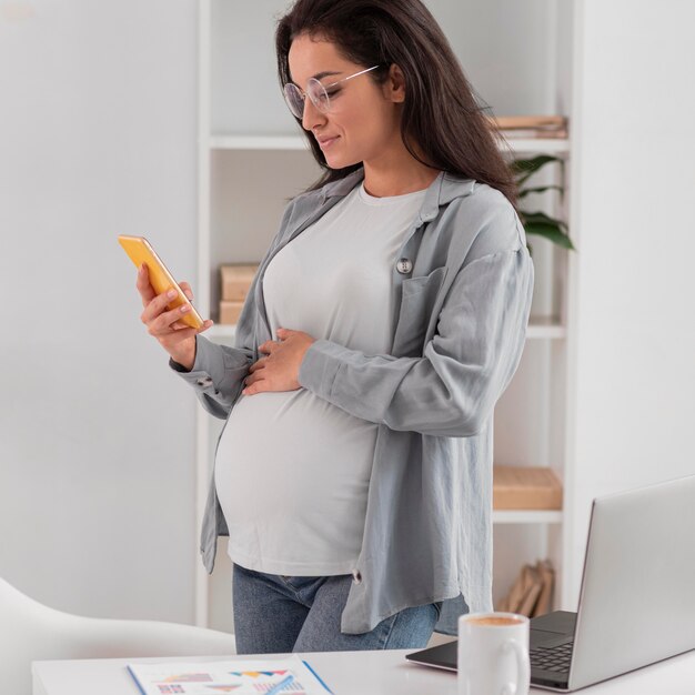 Vista lateral de la mujer embarazada en casa con ordenador portátil y smartphone
