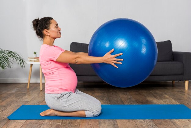 Vista lateral de la mujer embarazada haciendo ejercicio en la alfombra en casa con pelota