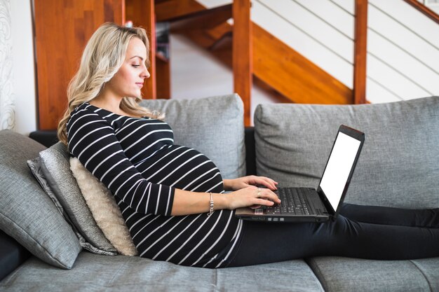 Vista lateral de una mujer embarazada que usa la computadora portátil con la pantalla blanca en blanco