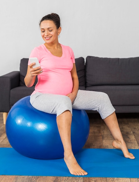 Vista lateral de la mujer embarazada sonriente en casa entrenando con pelota y con smartphone