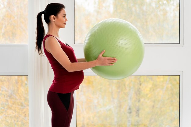 Vista lateral de la mujer embarazada sosteniendo una pelota verde fitness