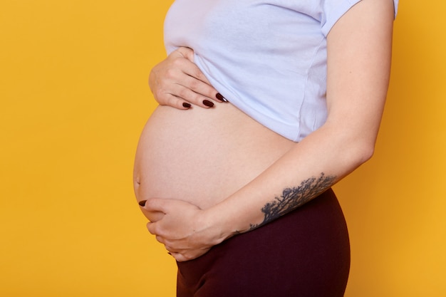 Vista lateral de la mujer embarazada con el vientre desnudo aislado sobre la pared amarilla. Mujer con tatuaje en hend vestida casualmente siendo fotografiada. Concepto de maternidad y embarazo.