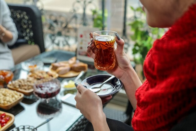 Vista lateral, una mujer está bebiendo té en un vaso de armudu con dulces sobre la mesa
