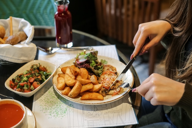 Vista lateral, una mujer está comiendo pollo monástico con papas fritas y ensalada de verduras con jugo sobre la mesa