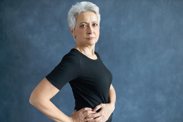 Vista lateral de una mujer europea de pelo gris serio con camiseta negra casual posando sobre fondo de pared de estudio de espacio de copia, tomados de la mano en la cintura, con expresión facial segura