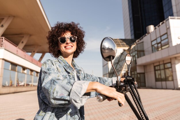 Vista lateral de la mujer feliz en gafas de sol posando en moto
