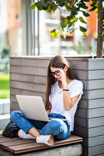 Vista lateral de una mujer joven en anteojos sentado en el banco en el parque y usando la computadora portátil