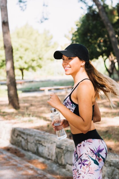 Vista lateral de una mujer joven de la aptitud que corre con la botella de agua en el parque