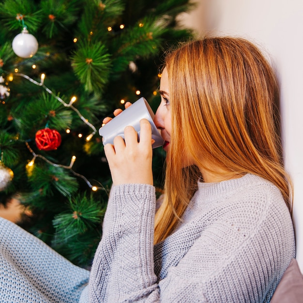 Vista lateral de mujer joven bebiendo té enfrente de árbol de navidad