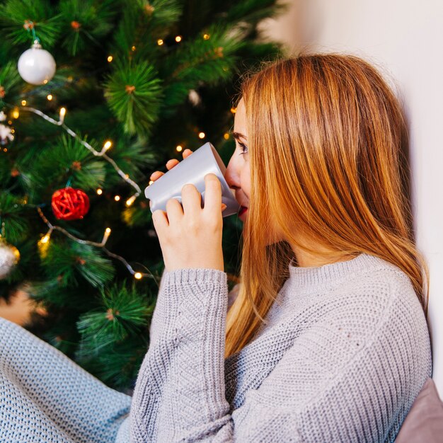 Vista lateral de mujer joven bebiendo té enfrente de árbol de navidad