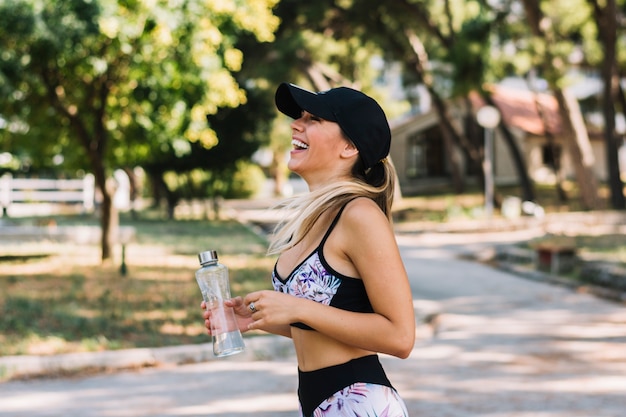 Vista lateral de una mujer joven feliz que se coloca en el parque que sostiene la botella de agua