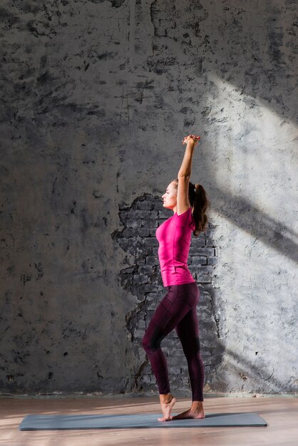 Vista lateral de una mujer joven que estira en la estera del ejercicio contra la pared gris vieja
