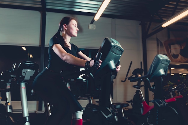 Vista lateral de una mujer joven que monta en la bicicleta estática en el gimnasio