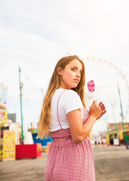 Vista lateral de la mujer joven que sostiene la piruleta grande en el parque de atracciones