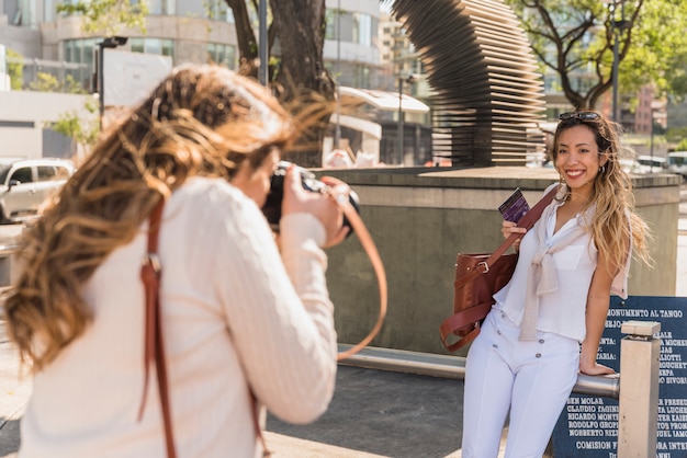 Foto gratuita vista lateral de una mujer joven que toma una foto de su amiga apoyada en una barandilla