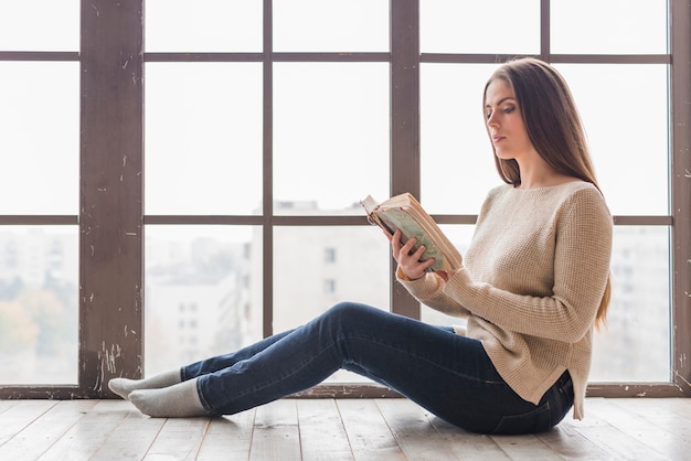 Vista lateral de una mujer joven sentada cerca del libro de lectura de la ventana