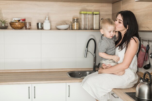 Vista lateral de una mujer joven sentada en el mostrador de la cocina amando a su hijo