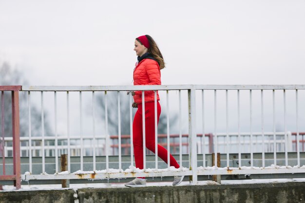 Vista lateral de la mujer joven sonriente que corre detrás de la barandilla en invierno