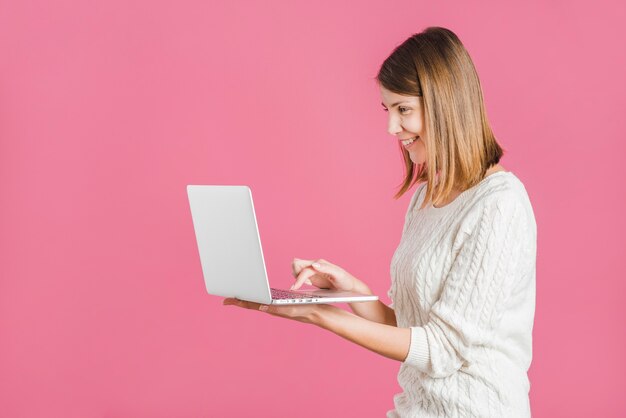 Vista lateral de una mujer joven sonriente que trabaja en la computadora portátil contra fondo rosado