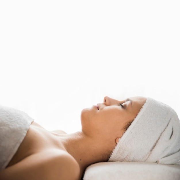 Vista lateral de la mujer joven con la toalla envuelta en su cabeza que duerme sobre cama del masaje contra el contexto blanco