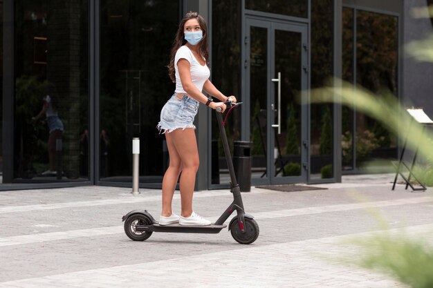 Vista lateral de la mujer con máscara montando un scooter eléctrico