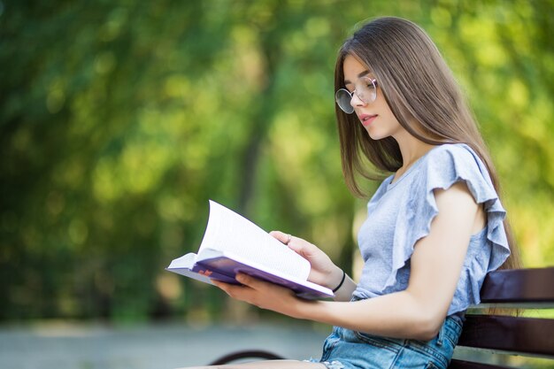 Vista lateral de la mujer morena complacida en anteojos sentado en un banco y libro de lectura en el parque