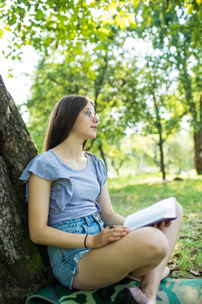 Vista lateral de la mujer morena complacida en anteojos sentado en la hierba bajo un árbol y libro de lectura en el parque