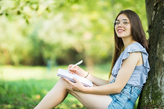 Vista lateral de la mujer morena concentrada en anteojos sentado cerca del árbol en el parque y escribiendo algo en el cuaderno