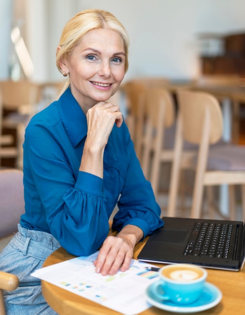 Vista lateral de la mujer de negocios anciana sonriente posando mientras trabaja en la computadora portátil y tomando café
